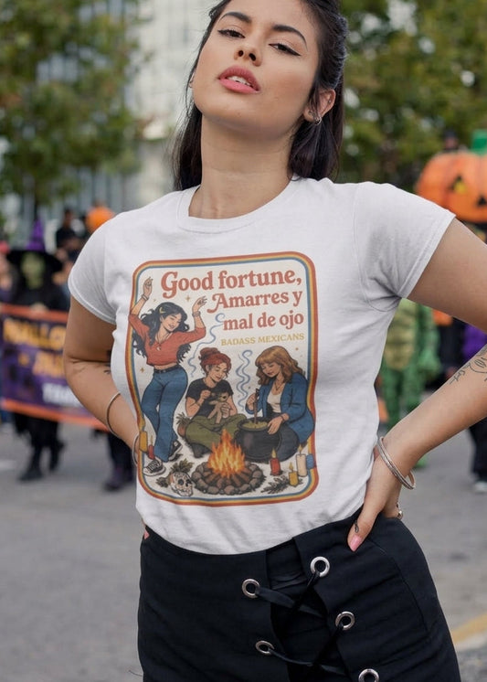White fitted t-shirt featuring a retro 70s illustration of three witches around a campfire with the text "Good fortune, Amarres y mal de ojo".