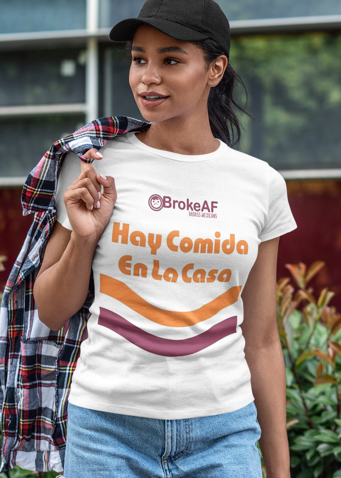 A woman wearing a white t-shirt featuring the phrase “Hay Comida En La Casa” in a retro instant noodle-inspired design with orange and brown accents.