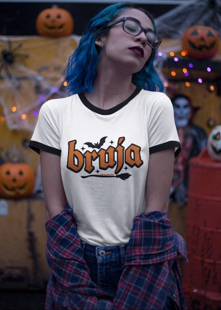 White ringer t-shirt with black collar and cuffs featuring the word "BRUJA" in bold orange font with a bat and broomstick graphic.