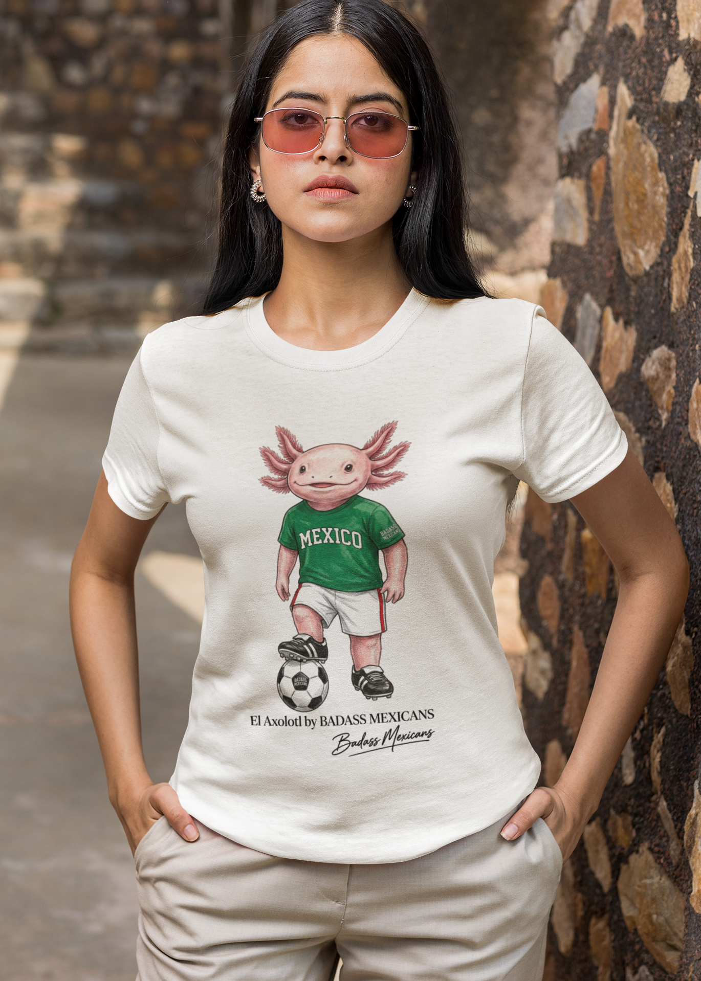 A white crew-neck t-shirt  featuring a colorful graphic illustration of a cute pink axolotl character dressed in a green Mexico soccer jersey, white shorts, and soccer cleats. The axolotl is standing with one foot on a classic black and white soccer ball. Below the design, it reads "El Axolotl by BADASS MEXICANS" with a cursive signature.