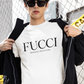 A white lifestyle t-shirt featuring the word "FUCCI" printed in a large, elegant black serif font across the chest. Below it, the brand name "BADASS MEXICANS" is printed in a smaller, clean sans-serif font.