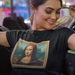A woman wearing a black unisex t-shirt, featuring a parody artwork inspired by the Mona Lisa portrayed with a chola aesthetic, including hoop earrings, a headband, and visible tattoos, framed like a classical painting.