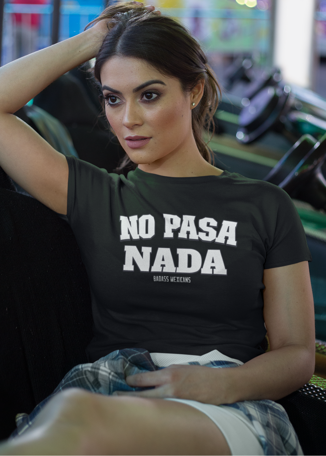 Woman wearing a black unisex t-shirt with the phrase “No Pasa Nada” printed in bold white letters, seated in an arcade environment with colorful lights in the background.