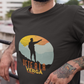 Black crew-neck t-shirt with a colorful retro circular sunset graphic showing a mountain hiker silhouette pointing forward above orange and yellow text "Vete a la verga".