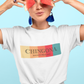A white fitted women's crew-neck t-shirt featuring a horizontal graphic design across the chest. The design features five vertical, vintage-toned color blocks lined up side-by-side in pastel yellow, soft orange, terracota red, deep pink, and muted teal. Overlaid across these colorful blocks, the word "CHINGONA" is printed in a large, elegant dark gray serif font, with the words "BADASS MEXICANS" written directly underneath in a smaller, clean sans-serif typeface.