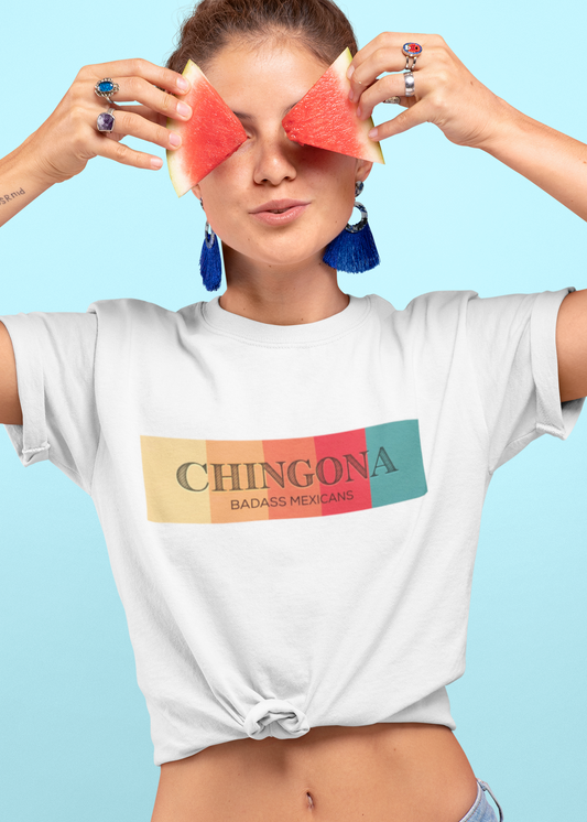 A white fitted women's crew-neck t-shirt featuring a horizontal graphic design across the chest. The design features five vertical, vintage-toned color blocks lined up side-by-side in pastel yellow, soft orange, terracota red, deep pink, and muted teal. Overlaid across these colorful blocks, the word "CHINGONA" is printed in a large, elegant dark gray serif font, with the words "BADASS MEXICANS" written directly underneath in a smaller, clean sans-serif typeface.