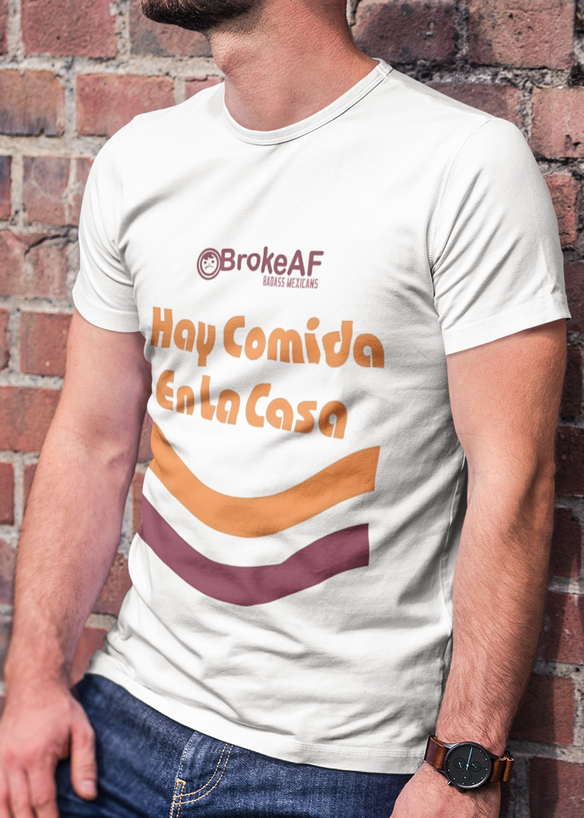 A man wearing a White t-shirt featuring the phrase “Hay Comida En La Casa” in a retro instant noodle-inspired design with orange and brown accents.
