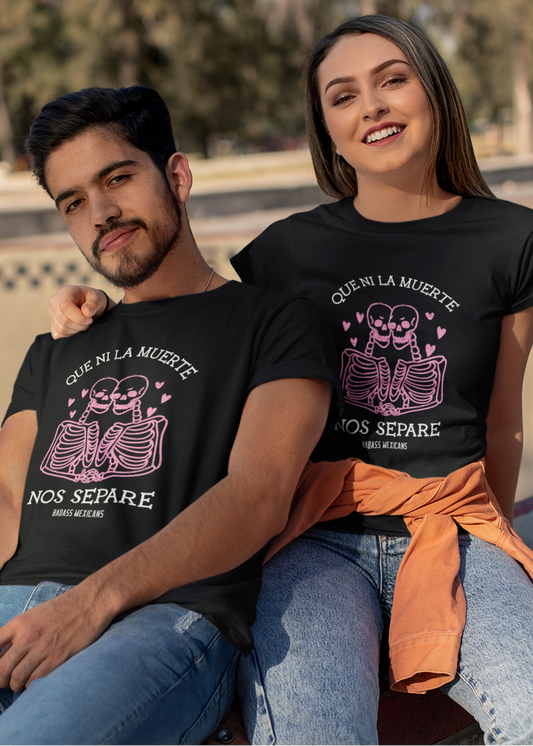 A couple sitting together at a skate park wearing matching black unisex t-shirts featuring a pink skeleton couple graphic with hearts and the phrase “Que Ni La Muerte Nos Separe,” photographed outdoors in daylight.