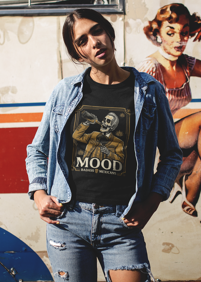 A high-quality graphic on a black t-shirt featuring a skeleton dressed in a traditional gold and brown Mariachi suit. The skeleton is holding a vintage microphone and drinking from a bottle. The design is framed in an elegant vintage border with the word "MOOD" in large serif letters and "BADASS MEXICANS" underneath.