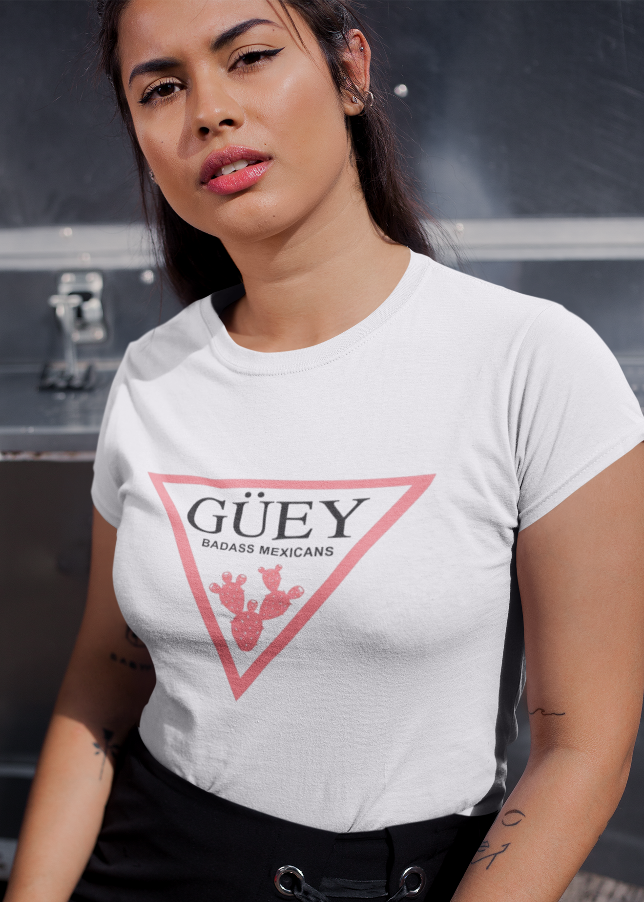 A white crew-neck t-shirt featuring a large, inverted pinkish-red triangle graphic printed on the chest. Inside the upper section of the triangle, the word "GÜEY" is written in a bold, black serif font, with the words "BADASS MEXICANS" printed right underneath in a smaller black sans-serif font. Centered at the bottom point of the triangle is a detailed graphic illustration of a prickly pear cactus (nopal) in a matching pinkish-red color.