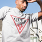 A white crew-neck t-shirt featuring a large, inverted pinkish-red triangle graphic printed on the chest. Inside the upper section of the triangle, the word "GÜEY" is written in a bold, black serif font, with the words "BADASS MEXICANS" printed right underneath in a smaller black sans-serif font. Centered at the bottom point of the triangle is a detailed graphic illustration of a prickly pear cactus (nopal) in a matching pinkish-red color.