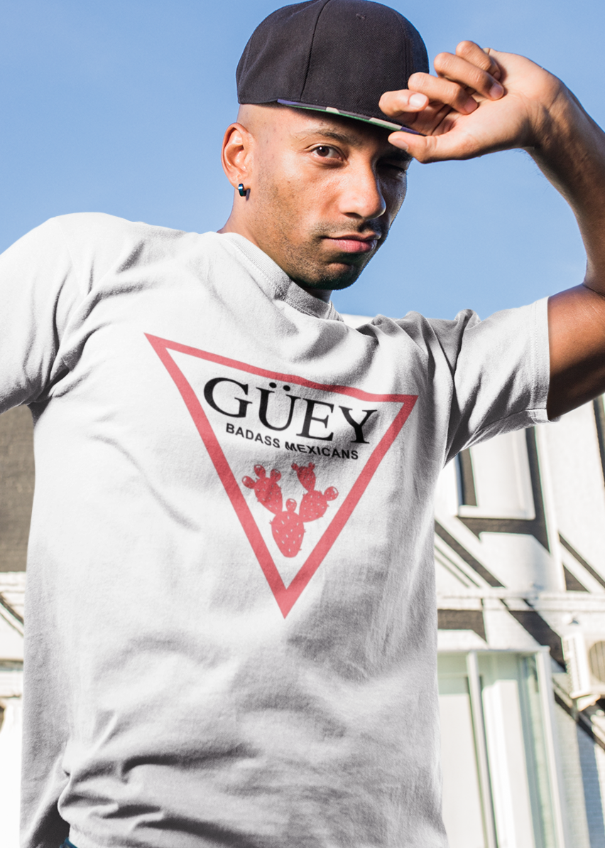 A white crew-neck t-shirt featuring a large, inverted pinkish-red triangle graphic printed on the chest. Inside the upper section of the triangle, the word "GÜEY" is written in a bold, black serif font, with the words "BADASS MEXICANS" printed right underneath in a smaller black sans-serif font. Centered at the bottom point of the triangle is a detailed graphic illustration of a prickly pear cactus (nopal) in a matching pinkish-red color.