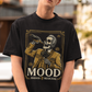 A high-quality graphic on a black t-shirt featuring a skeleton dressed in a traditional gold and brown Mariachi suit. The skeleton is holding a vintage microphone and drinking from a bottle. The design is framed in an elegant vintage border with the word "MOOD" in large serif letters and "BADASS MEXICANS" underneath.