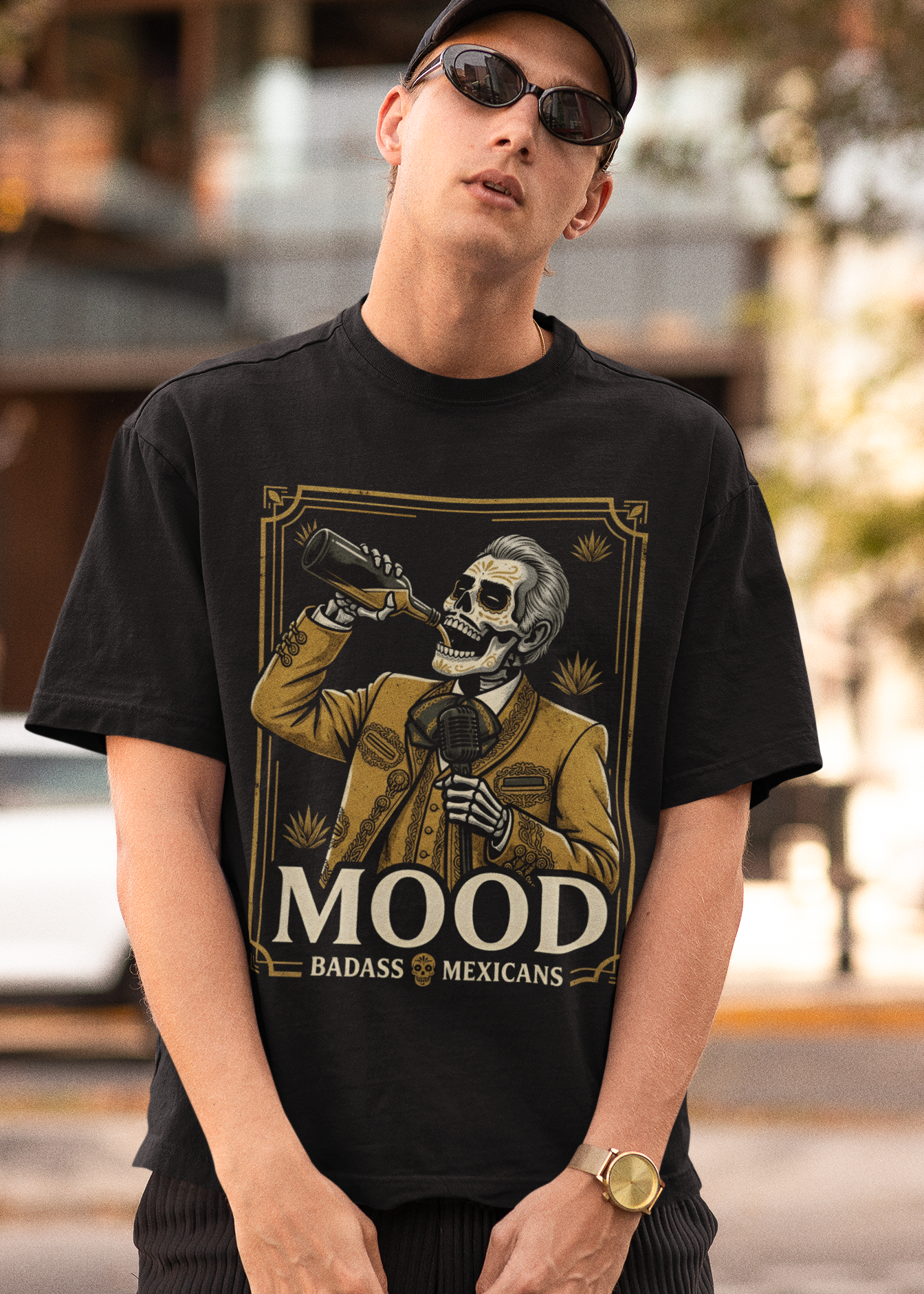 A high-quality graphic on a black t-shirt featuring a skeleton dressed in a traditional gold and brown Mariachi suit. The skeleton is holding a vintage microphone and drinking from a bottle. The design is framed in an elegant vintage border with the word "MOOD" in large serif letters and "BADASS MEXICANS" underneath.