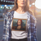 A woman wearing a white unisex t-shirt, featuring a parody artwork inspired by the Mona Lisa portrayed with a chola aesthetic, including hoop earrings, a headband, and visible tattoos, framed like a classical painting.
