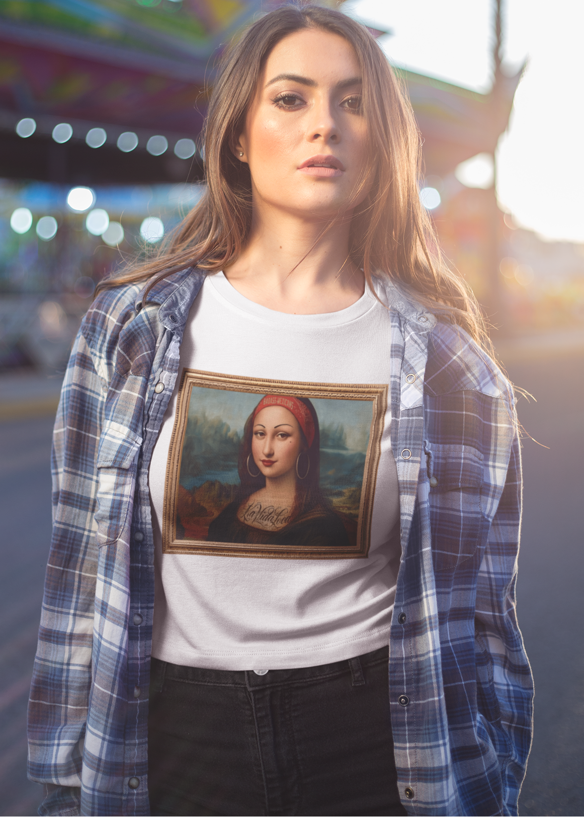 A woman wearing a white unisex t-shirt, featuring a parody artwork inspired by the Mona Lisa portrayed with a chola aesthetic, including hoop earrings, a headband, and visible tattoos, framed like a classical painting.