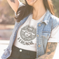 A white t-shirt featuring a large black graphic. The design includes a circular Aztec calendar-style background with a stylized man wearing a traditional Mexican charro hat in the center. The word "VERGACHI" is printed in a bold, arched serif font underneath the graphic, with "BADASS MEXICANS" in a small arch above it.
