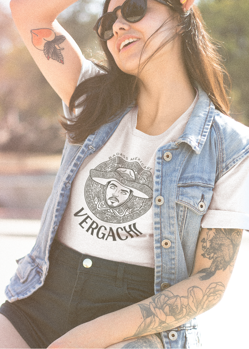 A white t-shirt featuring a large black graphic. The design includes a circular Aztec calendar-style background with a stylized man wearing a traditional Mexican charro hat in the center. The word "VERGACHI" is printed in a bold, arched serif font underneath the graphic, with "BADASS MEXICANS" in a small arch above it.