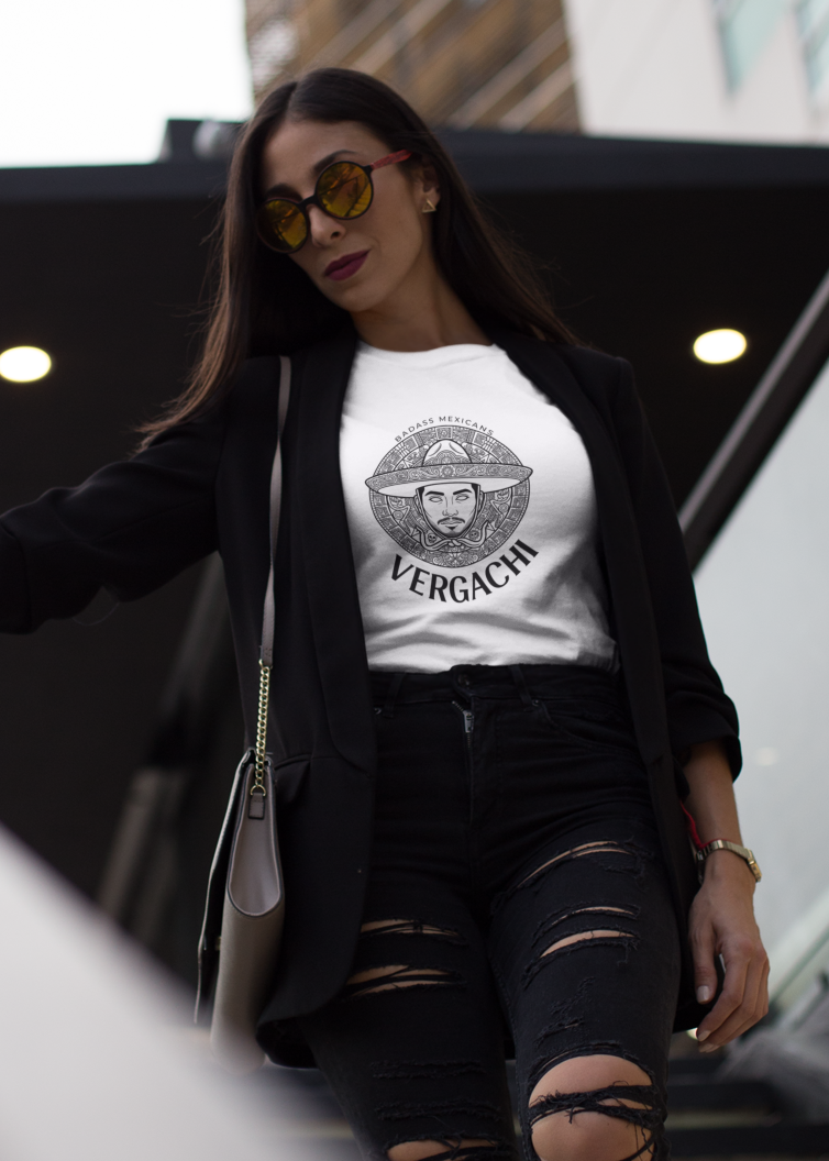 A white t-shirt featuring a large black graphic. The design includes a circular Aztec calendar-style background with a stylized man wearing a traditional Mexican charro hat in the center. The word "VERGACHI" is printed in a bold, arched serif font underneath the graphic, with "BADASS MEXICANS" in a small arch above it.