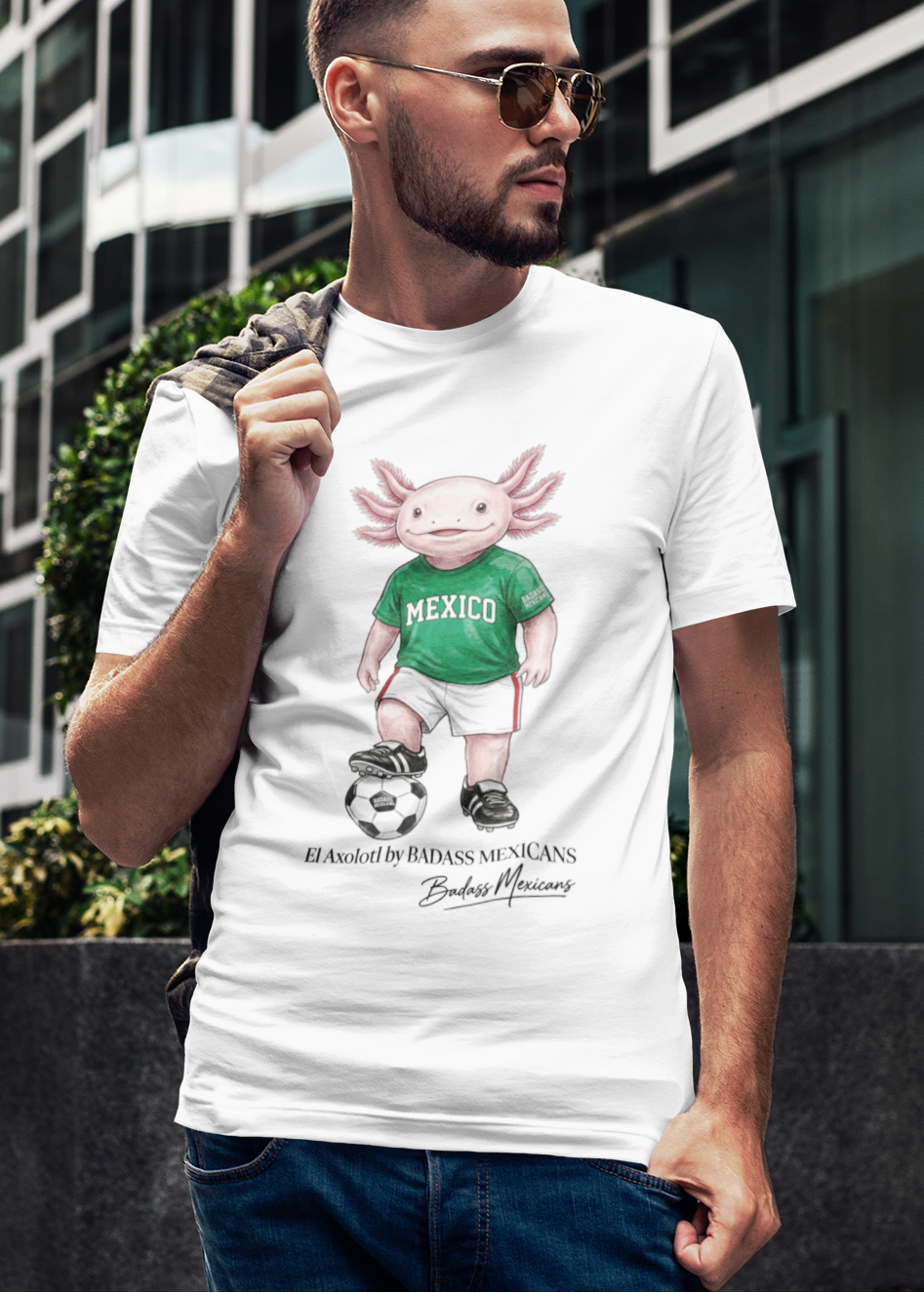 A white crew-neck t-shirt  featuring a colorful graphic illustration of a cute pink axolotl character dressed in a green Mexico soccer jersey, white shorts, and soccer cleats. The axolotl is standing with one foot on a classic black and white soccer ball. Below the design, it reads "El Axolotl by BADASS MEXICANS" with a cursive signature.