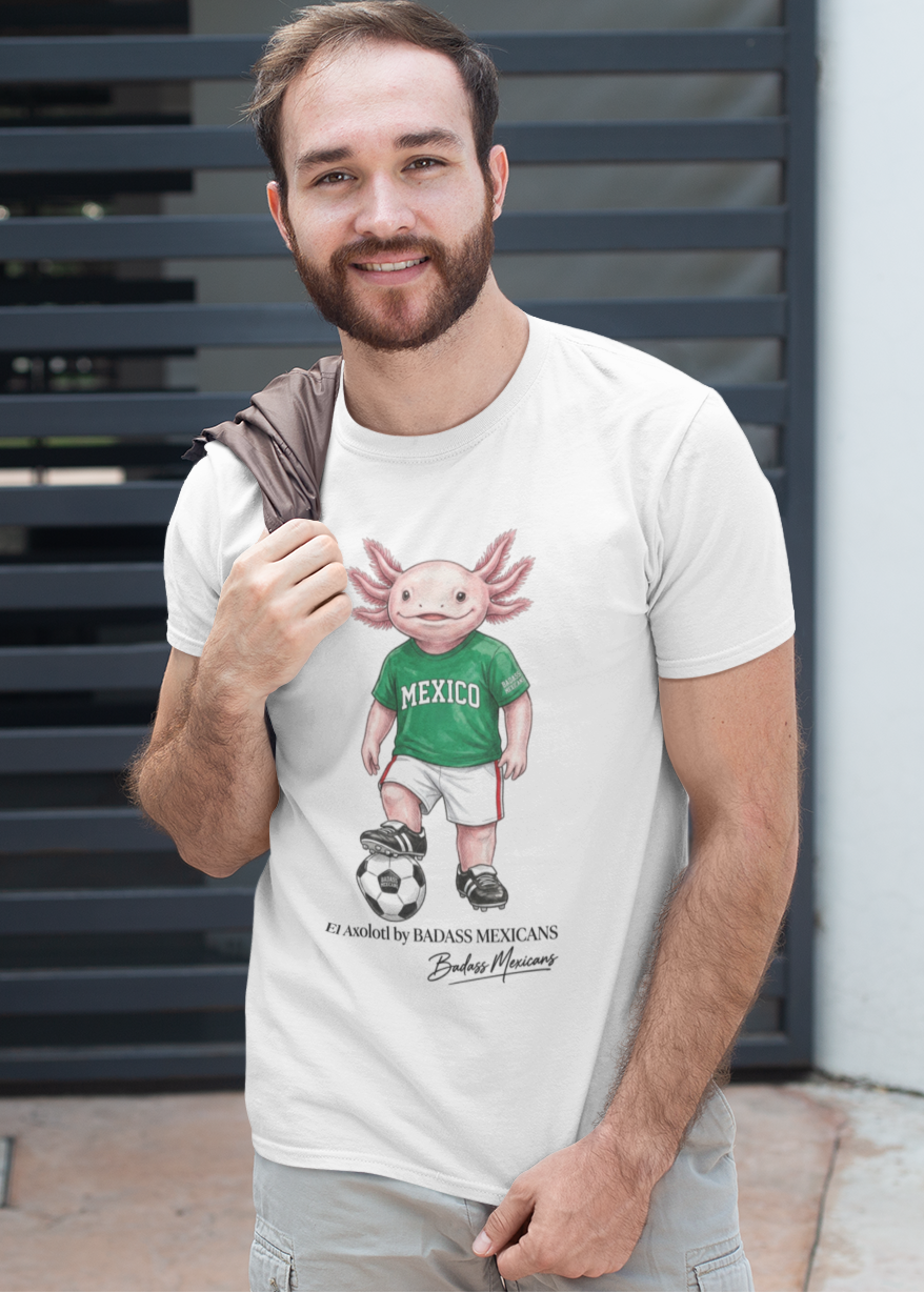 A white crew-neck t-shirt  featuring a colorful graphic illustration of a cute pink axolotl character dressed in a green Mexico soccer jersey, white shorts, and soccer cleats. The axolotl is standing with one foot on a classic black and white soccer ball. Below the design, it reads "El Axolotl by BADASS MEXICANS" with a cursive signature.