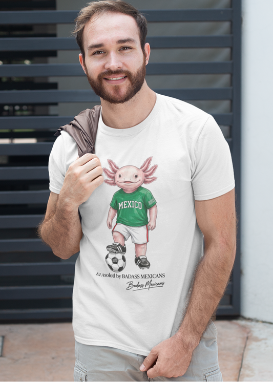 A white crew-neck t-shirt  featuring a colorful graphic illustration of a cute pink axolotl character dressed in a green Mexico soccer jersey, white shorts, and soccer cleats. The axolotl is standing with one foot on a classic black and white soccer ball. Below the design, it reads "El Axolotl by BADASS MEXICANS" with a cursive signature.