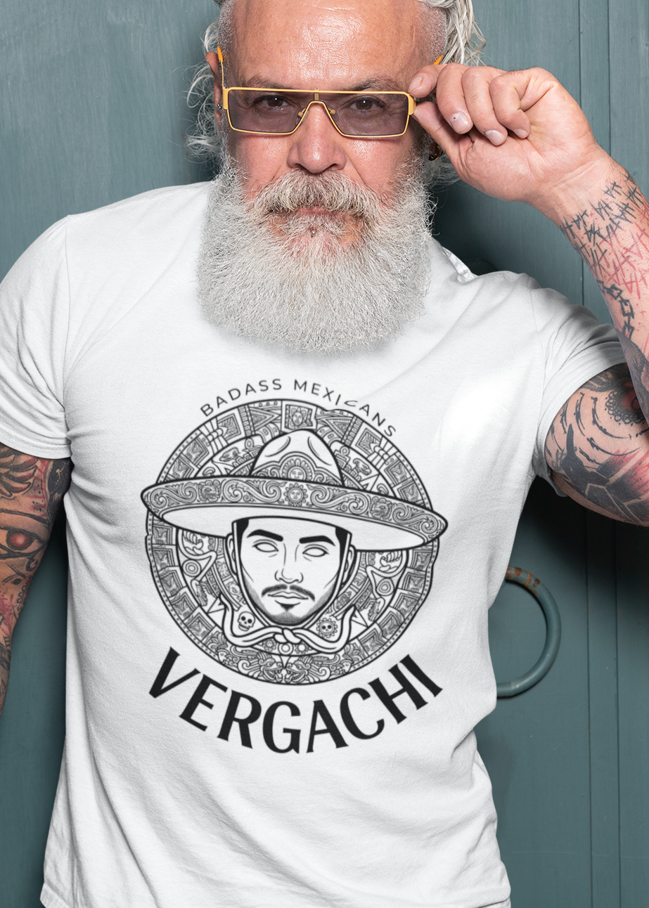 A white t-shirt featuring a large black graphic. The design includes a circular Aztec calendar-style background with a stylized man wearing a traditional Mexican charro hat in the center. The word "VERGACHI" is printed in a bold, arched serif font underneath the graphic, with "BADASS MEXICANS" in a small arch above it.