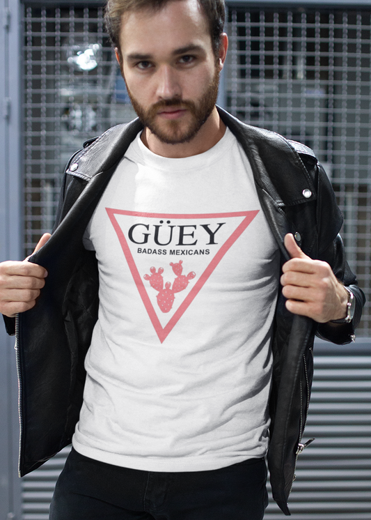 A white crew-neck t-shirt featuring a large, inverted pinkish-red triangle graphic printed on the chest. Inside the upper section of the triangle, the word "GÜEY" is written in a bold, black serif font, with the words "BADASS MEXICANS" printed right underneath in a smaller black sans-serif font. Centered at the bottom point of the triangle is a detailed graphic illustration of a prickly pear cactus (nopal) in a matching pinkish-red color.