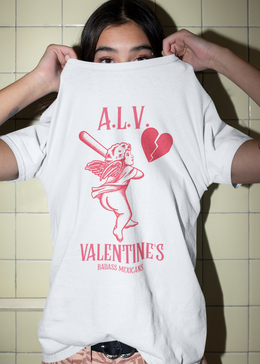 A woman holding up a white unisex t-shirt that features a red cherub graphic holding a bat, a broken heart icon, and the text “A.L.V. Valentine’s Badass Mexicans,” inspired by sarcastic Mexican Valentine’s Day humor.