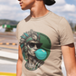 A sand crew-neck t-shirt featuring a circular graphic illustration on the chest. The design depicts an Aztec ruler or warrior chief in profile, wearing a highly detailed traditional feathered headdress and ear spools in shades of turquoise, green, and gray. The character is styled with a modern twist, wearing black sunglasses and blowing a large, bright light-blue bubblegum bubble. The entire illustration is set against a dark green solid circular background with the words "BADASS MEXICANS" written.