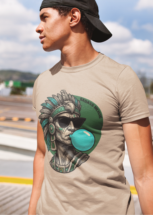 A sand crew-neck t-shirt featuring a circular graphic illustration on the chest. The design depicts an Aztec ruler or warrior chief in profile, wearing a highly detailed traditional feathered headdress and ear spools in shades of turquoise, green, and gray. The character is styled with a modern twist, wearing black sunglasses and blowing a large, bright light-blue bubblegum bubble. The entire illustration is set against a dark green solid circular background with the words "BADASS MEXICANS" written.