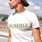 A white streetwear t-shirt showcasing the word "BALENBERGA" across the chest in a heavy, textured, geometric block font with alternating mirrored letters in an olive-beige tone. Underneath, "BADASS MEXICANS" is printed in a clean, sharp matching font.