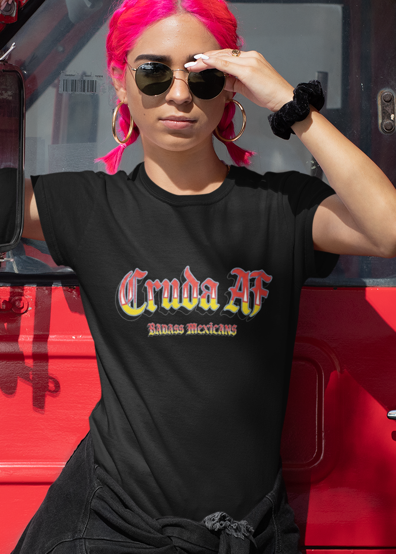 A black fitted women's crew-neck t-shirt featuring the text "Cruda AF" printed across the chest in a bold, stylized Gothic font. The letters feature a horizontal color gradient transition from red at the top, fading into yellow and white at the bottom, outlined in black with a heavy black drop shadow. Directly below, the words "BADASS MEXICANS" are printed in a smaller, matching Gothic font with the same red, yellow, and white gradient and drop shadow effect.