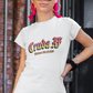 A white fitted women's crew-neck t-shirt featuring the text "Cruda AF" printed across the chest in a bold, stylized Gothic font. The letters feature a horizontal color gradient transition from red at the top, fading into yellow and white at the bottom, outlined in black with a heavy black drop shadow. Directly below, the words "BADASS MEXICANS" are printed in a smaller, matching Gothic font with the same red, yellow, and white gradient and drop shadow effect.