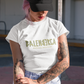A white streetwear t-shirt showcasing the word "BALENBERGA" across the chest in a heavy, textured, geometric block font with alternating mirrored letters in an olive-beige tone. Underneath, "BADASS MEXICANS" is printed in a clean, sharp matching font.
