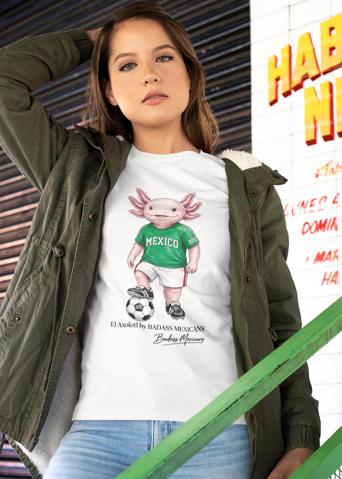 A white crew-neck t-shirt  featuring a colorful graphic illustration of a cute pink axolotl character dressed in a green Mexico soccer jersey, white shorts, and soccer cleats. The axolotl is standing with one foot on a classic black and white soccer ball. Below the design, it reads "El Axolotl by BADASS MEXICANS" with a cursive signature.