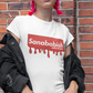 A white crew-neck t-shirt featuring a solid red rectangular box graphic on the chest. Inside the box, the word "Sanababish" is printed in a bold white, slanted font, with the words "BADASS MEXICANS" in smaller white letters beneath it. The bottom edge of the red box is styled to look like wet paint, with thick red droplets dripping down onto the fabric.