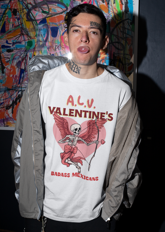 A person wearing a white unisex t-shirt with a skeleton cupid graphic holding a bow and arrow in front of a pink heart, with the text “A.L.V. Valentine’s Badass Mexicans,” photographed indoors against a colorful abstract painting background.