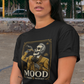 A high-quality graphic on a black t-shirt featuring a skeleton dressed in a traditional gold and brown Mariachi suit. The skeleton is holding a vintage microphone and drinking from a bottle. The design is framed in an elegant vintage border with the word "MOOD" in large serif letters and "BADASS MEXICANS" underneath.