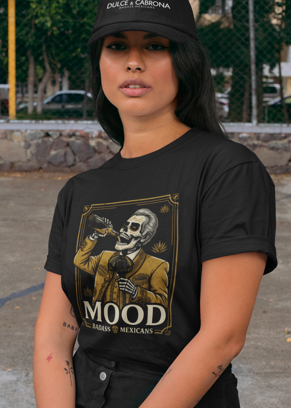A high-quality graphic on a black t-shirt featuring a skeleton dressed in a traditional gold and brown Mariachi suit. The skeleton is holding a vintage microphone and drinking from a bottle. The design is framed in an elegant vintage border with the word "MOOD" in large serif letters and "BADASS MEXICANS" underneath.
