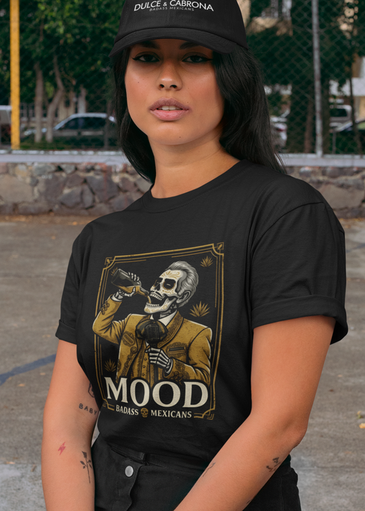 A high-quality graphic on a black t-shirt featuring a skeleton dressed in a traditional gold and brown Mariachi suit. The skeleton is holding a vintage microphone and drinking from a bottle. The design is framed in an elegant vintage border with the word "MOOD" in large serif letters and "BADASS MEXICANS" underneath.