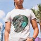 A white crew-neck t-shirt featuring a circular graphic illustration on the chest. The design depicts an Aztec ruler or warrior chief in profile, wearing a highly detailed traditional feathered headdress and ear spools in shades of turquoise, green, and gray. The character is styled with a modern twist, wearing black sunglasses and blowing a large, bright light-blue bubblegum bubble. The entire illustration is set against a dark green solid circular background with the words "BADASS MEXICANS" written