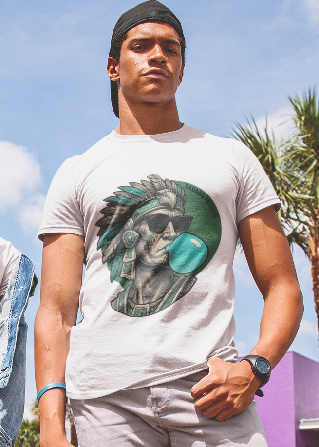 A white crew-neck t-shirt featuring a circular graphic illustration on the chest. The design depicts an Aztec ruler or warrior chief in profile, wearing a highly detailed traditional feathered headdress and ear spools in shades of turquoise, green, and gray. The character is styled with a modern twist, wearing black sunglasses and blowing a large, bright light-blue bubblegum bubble. The entire illustration is set against a dark green solid circular background with the words "BADASS MEXICANS" written