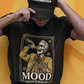 A high-quality graphic on a black t-shirt featuring a skeleton dressed in a traditional gold and brown Mariachi suit. The skeleton is holding a vintage microphone and drinking from a bottle. The design is framed in an elegant vintage border with the word "MOOD" in large serif letters and "BADASS MEXICANS" underneath.