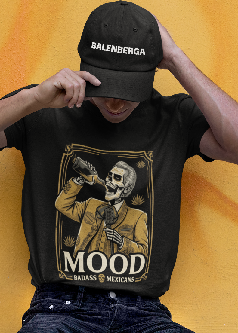 A high-quality graphic on a black t-shirt featuring a skeleton dressed in a traditional gold and brown Mariachi suit. The skeleton is holding a vintage microphone and drinking from a bottle. The design is framed in an elegant vintage border with the word "MOOD" in large serif letters and "BADASS MEXICANS" underneath.