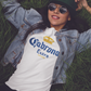 A woman wearing a White unisex t-shirt featuring the phrase “Cabrona Extra” in bold blue lettering with a gold crown above, inspired by a classic beer-style typography.