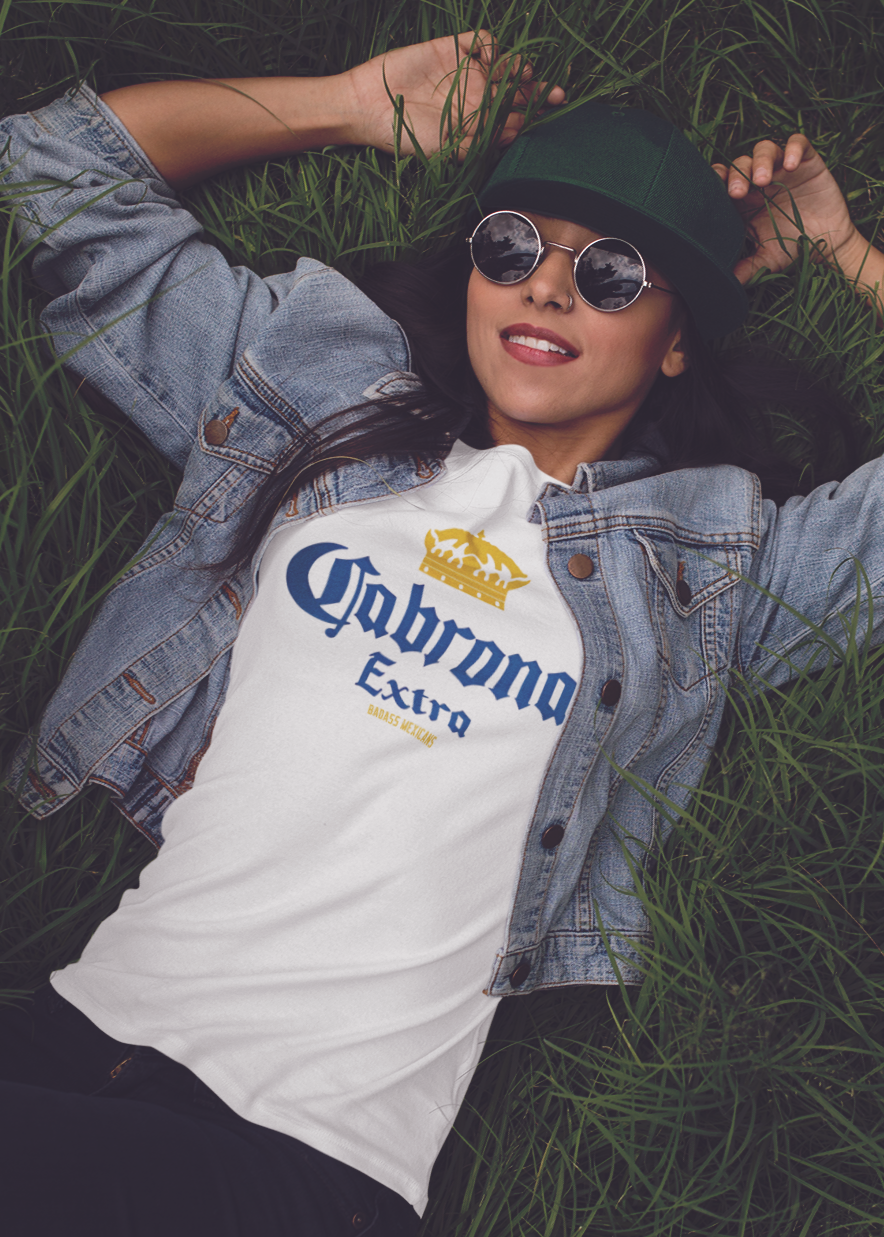 A woman wearing a White unisex t-shirt featuring the phrase “Cabrona Extra” in bold blue lettering with a gold crown above, inspired by a classic beer-style typography.