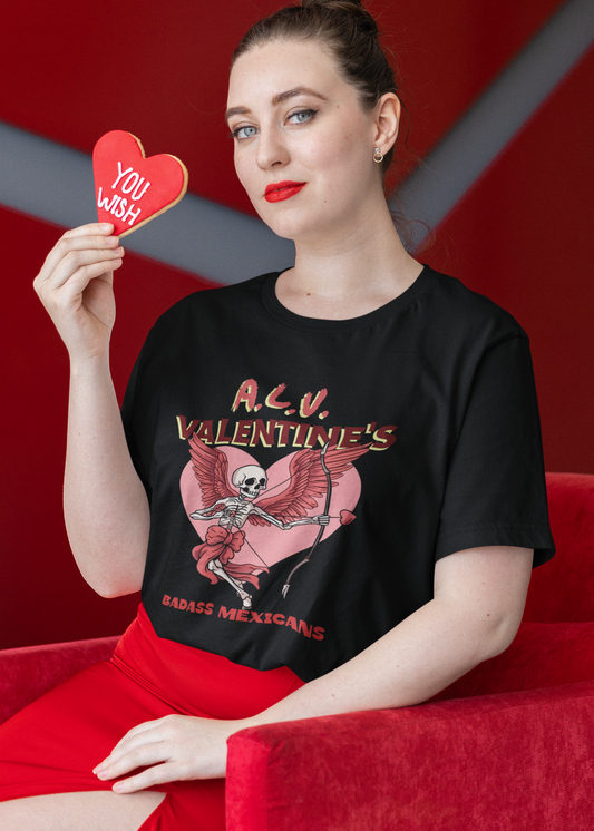 A person wearing a black unisex t-shirt with a skeleton cupid graphic holding a bow and arrow in front of a pink heart, with the text “A.L.V. Valentine’s Badass Mexicans,” 
